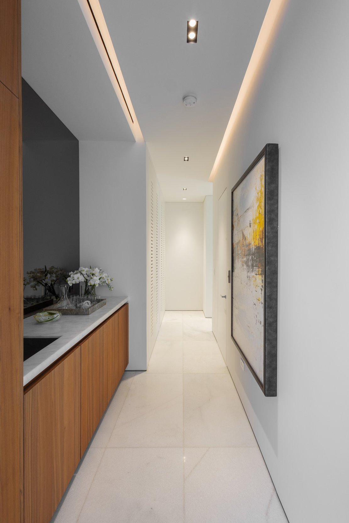 Modern hallway design featuring custom wood cabinetry with a marble countertop, recessed cove lighting, and minimalist white walls.