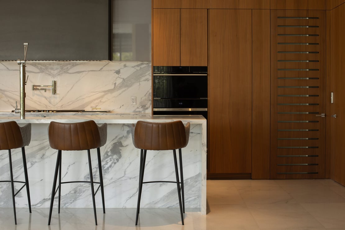 Custom kitchen featuring warm wood vertical cabinetry, an integrated black wall oven, and a marble breakfast bar with leather stools.