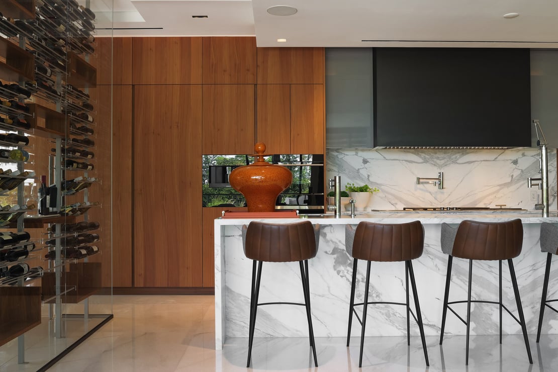 Armonia Materica kitchen featuring a marble waterfall island with leather barstools, warm wood cabinetry, and a custom glass-enclosed wine cellar.