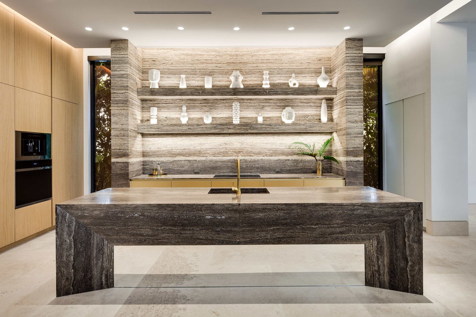 Modern luxury kitchen featuring a monolithic dark travertine island with a gold faucet, matching backlit stone floating shelves, and light wood custom cabinetry.