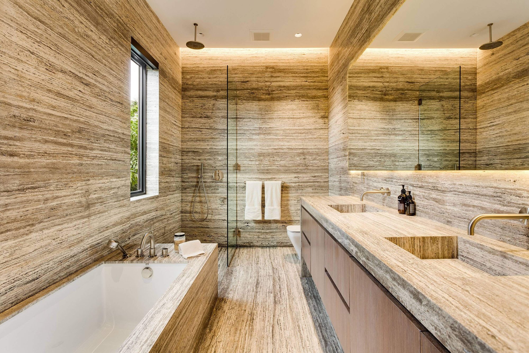 Immersive luxury bathroom entirely clad in veined light travertine stone floors and walls, featuring matching custom stone vanities with wood cabinetry, an integrated stone bathtub, and brass fixtures.