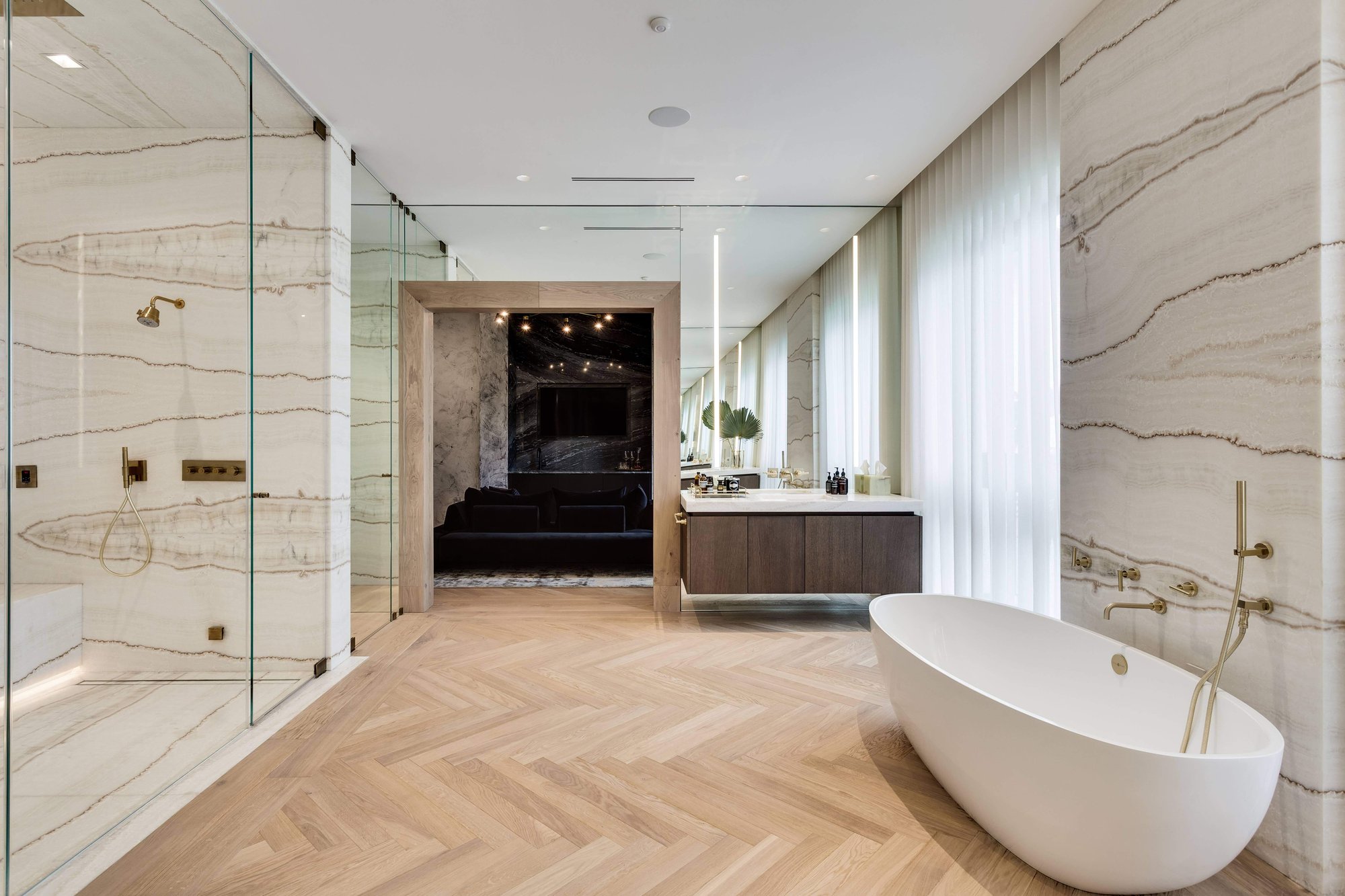 Immersive luxury bathroom with light wood herringbone flooring, bookmatched marble walls and glass-encased shower, a floating dark wood vanity, and a freestanding oval soaking tub with brass fixtures.