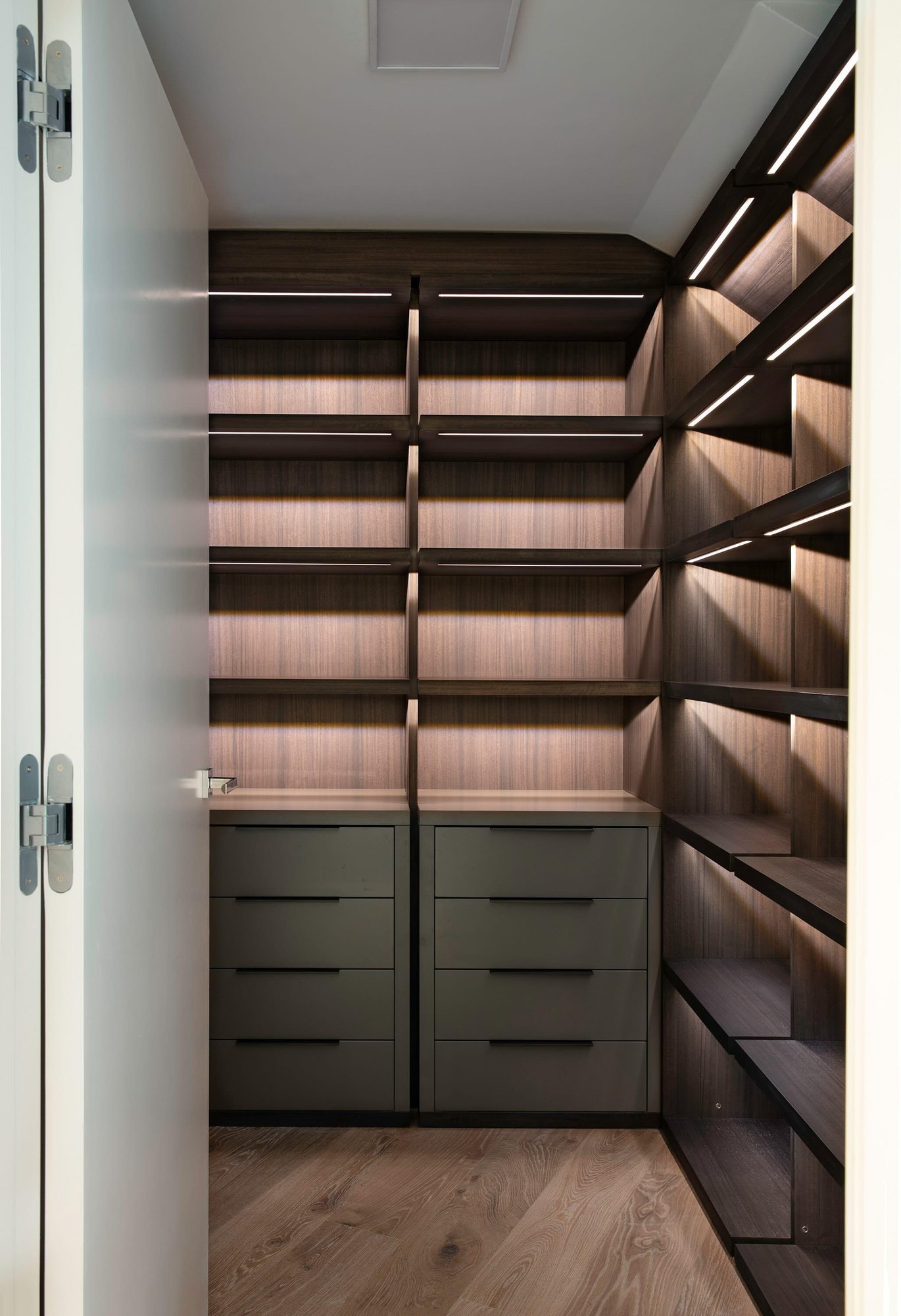 Modern dark wood walk-in closet with integrated LED shelf lighting and grey drawers.
