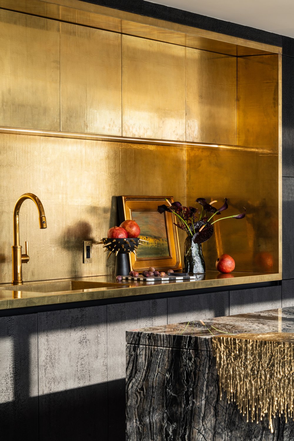 Luxury kitchen featuring a textured gold metal backsplash and recessed upper cabinetry, a gold faucet and sink, and a dark marble island with unique gold fringe detailing.