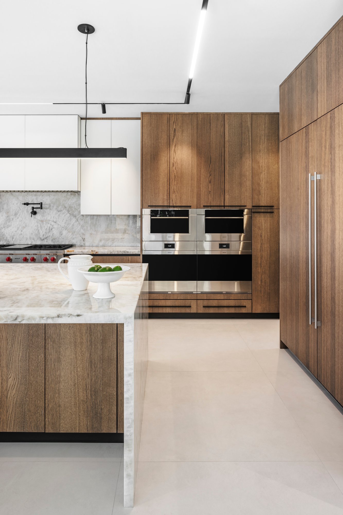 Luxury kitchen featuring a white marble waterfall island, natural wood grain cabinetry, and integrated Wolf wall ovens with a marble backsplash.
