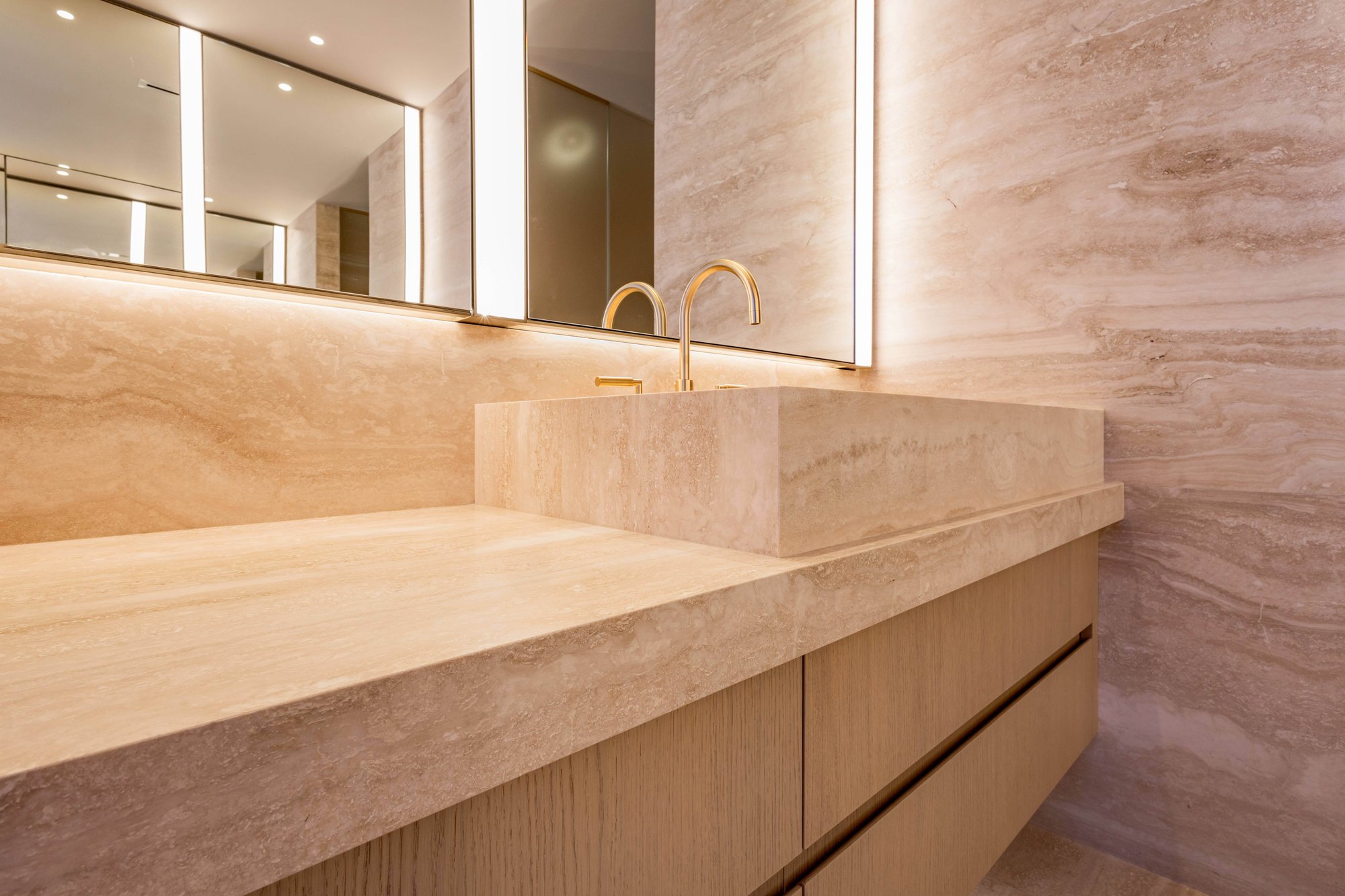 Detailed view of a luxury bathroom featuring an integrated travertine stone vessel sink on a stone countertop, warm natural wood cabinetry, matching travertine walls, and backlit mirrors with a gold faucet.