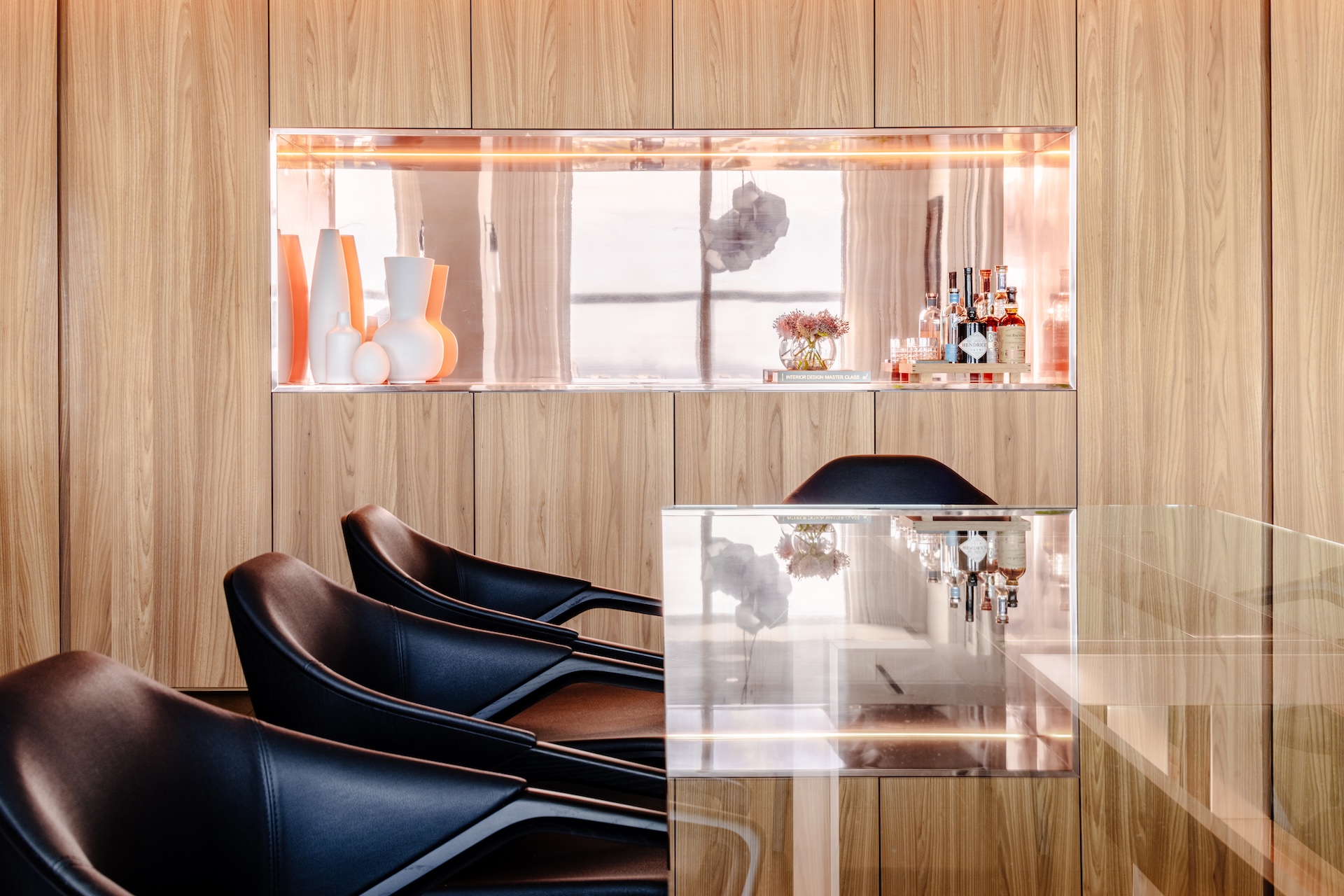 Warm wood-paneled interior with black chairs, illuminated display niche, and glass table in a modern luxury space.